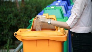 노란색 플라스틱 재활용 쓰레기통에 플라스틱 물병을 넣는 사람. 이 쓰레기통은 여러 다른 파란색 및 녹색 쓰레기통으로 구성되어 있습니다.